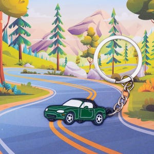 May include: A green car keychain with a silver key ring. The keychain is shaped like a convertible sports car with a black roof. The keychain is on a winding road with green trees and mountains in the background.