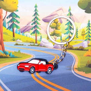 Puede incluir: Un llavero de coche rojo con techo negro y un llavero plateado. El llavero está en un camino sinuoso con árboles verdes y un cielo azul de fondo.