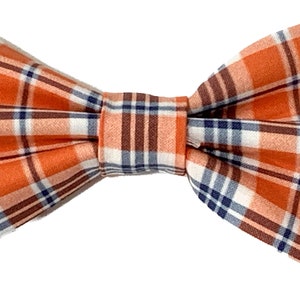 May include: A plaid bow tie in shades of orange, brown, white, and navy blue. The bow tie features a classic plaid pattern, suitable for formal or casual wear. The fabric appears to be a woven material.