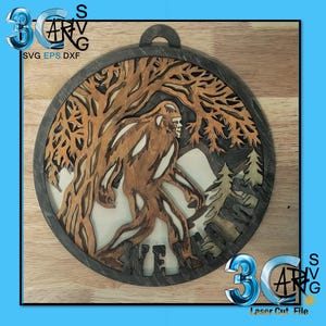 May include: A wooden Bigfoot-themed wall hanging. The design features a detailed silhouette of Bigfoot walking through a forest, with trees and mountains. The artwork is laser cut and has a dark gray border and a hanging loop.
