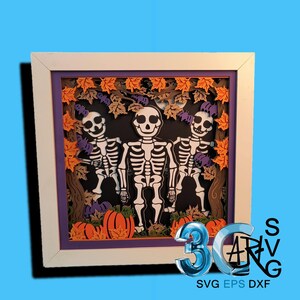 May include: A framed Halloween-themed 3D paper art piece. The artwork features three white skeletons standing in front of a black background with orange pumpkins, purple bats, and autumn leaves. The frame is white with a purple border.