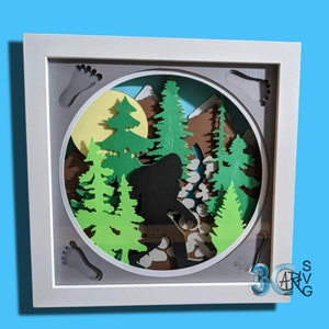 May include: A white frame with a gray mat surrounding a paper cut-out scene of a Bigfoot walking through a forest. The scene is set against a yellow moon and includes brown mountains, green trees, and gray footprints.