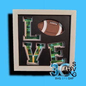 May include: A framed artwork with the word "LOVE" in green and white, with a football graphic. The letters are layered to create a 3D effect, with a football field design inside. The frame is white, and the background is black.