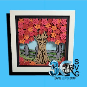 May include: A framed paper art piece featuring a layered autumn scene. The artwork depicts a tree with red and orange leaves, a brown trunk, and a wooden fence. The frame is white, and the background is a light blue.