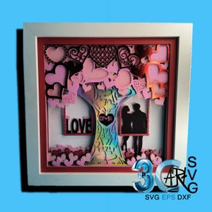 May include: A white frame with a red mat surrounding a layered paper cut design of a tree with hearts, a couple on a swing, and the word "LOVE".