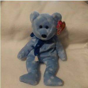 Op de afbeelding: Een blauwe en witte beanie baby beer met een patroon, die een blauwe sjaal met een witte kwast draagt. De beer heeft zwarte ogen en een zwarte neus. De beer heeft een rood en wit Ty-label aan zijn rechteroor.