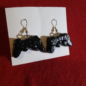 May include: A pair of black video game controller earrings. The earrings are made of resin and have silver hooks.