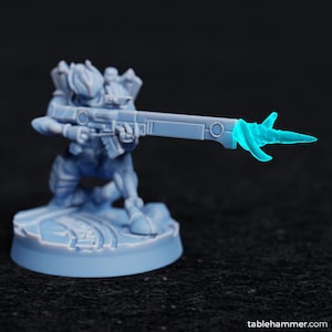 May include: A blue plastic miniature figure of a humanoid holding a rifle with a glowing blue energy effect at the end of the barrel. The figure is standing on a round base.