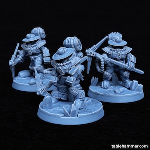 May include: Three blue and gray miniature figures of robots wearing helmets and holding tools. The figures are standing on round bases with a rocky texture.