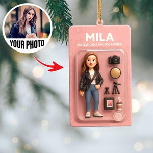 May include: A pink Christmas ornament featuring a cartoon figure of a photographer named Mila. The figure is wearing a black jacket and jeans. The ornament includes a camera, tripod, and coffee cup. The text "YOUR PHOTO" is also visible.