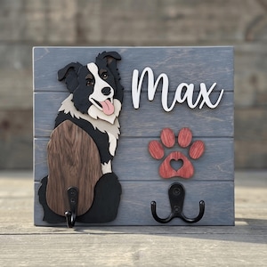 May include: A gray wooden wall-mounted hook rack featuring a layered cutout of a black and white Border Collie dog, the name Max in white script, a red wooden paw print with a heart cutout, and two black metal hooks for hanging items.