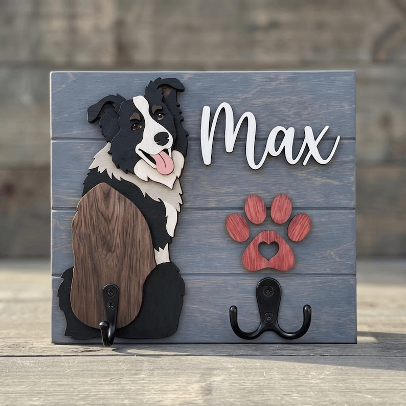 May include: A gray wooden wall-mounted hook rack featuring a layered cutout of a black and white Border Collie dog, the name Max in white script, a red wooden paw print with a heart cutout, and two black metal hooks for hanging items.
