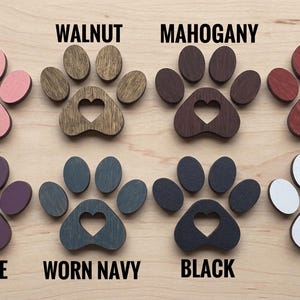 May include: Eight wooden paw print cutouts with heart-shaped centers, arranged in two rows. Colors include pink, walnut, mahogany, red, purple, worn navy, black, and white. Each paw print is labeled with its corresponding color name in black text.