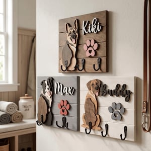 May include: Three rectangular wooden wall-mounted organizers featuring layered dog silhouettes, paw prints, and names. The top sign is brown with Kiki, the bottom left is gray with Max, and the bottom right is white with Buddy. Each has three black hooks.