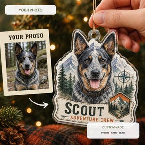 May include: A custom acrylic Christmas ornament featuring a portrait of a blue heeler dog, a mountain landscape, a compass, and the text SCOUT ADVENTURE CREW. The ornament includes a space for a personal photo and is suspended by a gold string.