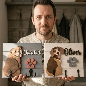 May include: Two rectangular wooden wall plaques featuring 3D dog cutouts. The left plaque is gray with a bulldog, the name Churchill, a red paw print, and two black hooks. The right plaque is white with a dachshund, the name Oliver, a gray paw print, and two black hooks.