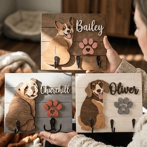 May include: Three rectangular wooden wall-mounted leash holders featuring layered wood cutouts of a Corgi, a Bulldog, and a Dachshund. Each board displays a dog name, a paw print cutout, and black metal hooks for hanging items on the wall.