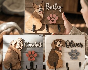 May include: Three rectangular wooden wall-mounted leash holders featuring layered wood cutouts of a Corgi, a Bulldog, and a Dachshund. Each board displays a dog name, a paw print cutout, and black metal hooks for hanging items on the wall.
