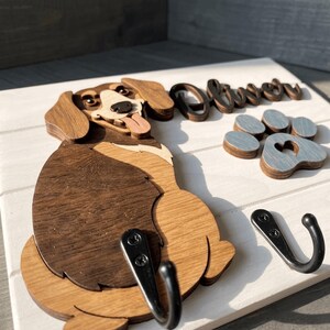 May include: A white wooden wall hook featuring a layered wooden dog cutout in shades of brown, a gray wooden paw print with a heart cutout, and the name Oliver in dark brown script. Two black metal hooks are attached to the base for hanging items.