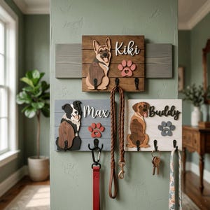 May include: Three wooden wall-mounted pet leash holders featuring cutouts of a German Shepherd, a Border Collie, and a mixed-breed dog. Each board displays a name, Kiki, Max, or Buddy, with a matching paw print and black metal hooks for leashes.