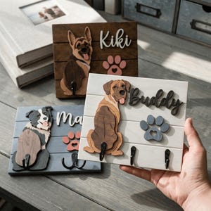 May include: Three rectangular wooden wall-mounted key holders featuring layered dog cutouts, names in script lettering, and paw print accents. Each board includes black metal hooks for hanging items, with one white board labeled Buddy.