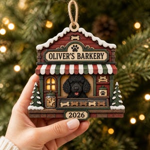 May include: A wooden Christmas tree ornament shaped like a shop titled OLIVER'S BARKERY, featuring a black poodle, a red and green striped awning, a jar of bone-shaped treats, two small pine trees with snow, and the year 2026 on a bottom plaque.