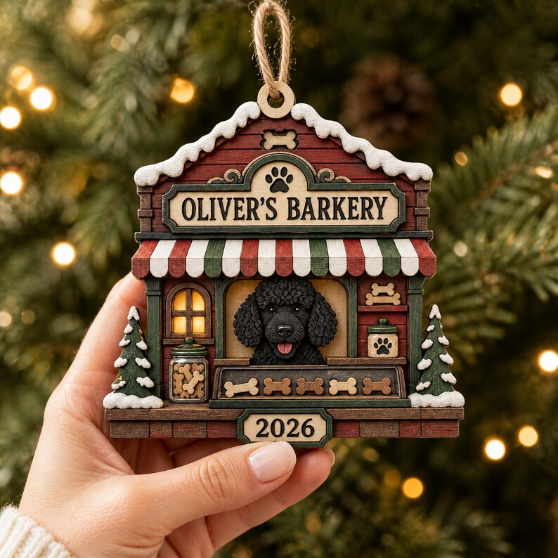 May include: A wooden Christmas tree ornament shaped like a shop titled OLIVER'S BARKERY, featuring a black poodle, a red and green striped awning, a jar of bone-shaped treats, two small pine trees with snow, and the year 2026 on a bottom plaque.