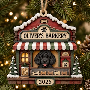 May include: A wooden Christmas tree ornament shaped like a shop titled OLIVER'S BARKERY, featuring a black poodle, a red and green striped awning, a jar of bone-shaped treats, and the year 2026. The structure has snow-dusted roof edges.