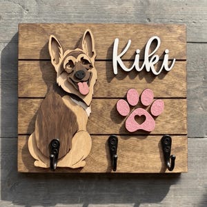 May include: A rectangular wooden wall-mounted hook rack featuring a layered German Shepherd cutout, a pink paw print with a heart cutout, and the white script text Kiki. Three black metal hooks are attached to the stained wood base.