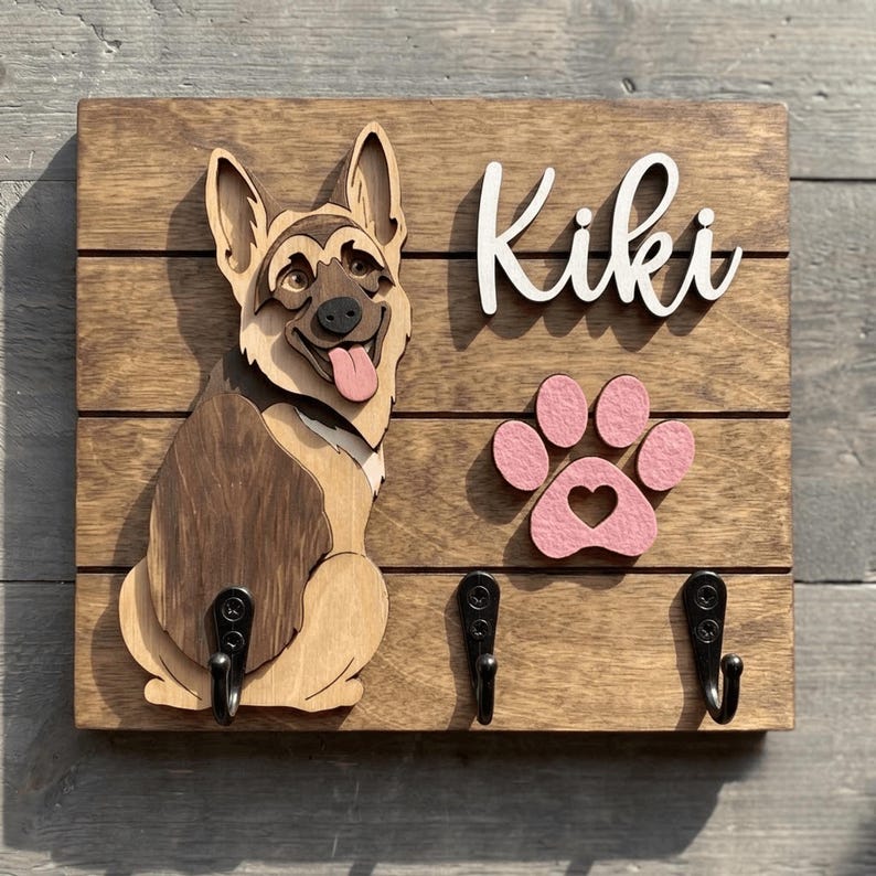May include: A rectangular wooden wall-mounted hook rack featuring a layered German Shepherd cutout, a pink paw print with a heart cutout, and the white script text Kiki. Three black metal hooks are attached to the stained wood base.