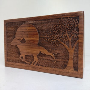 May include: A wooden box with a carved design of a horse running through a moonlit forest. The horse is silhouetted against a full moon, and a tree with leaves is in the background.