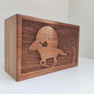 May include: A wooden box with a carved design of a person riding a horse in front of a full moon and a tree. The box is made of dark wood with a natural grain.