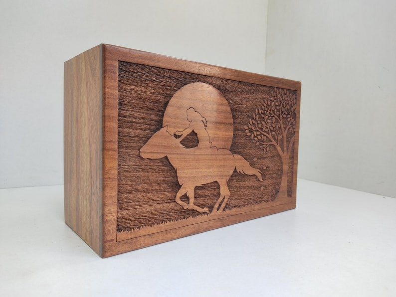 May include: A wooden box with a carved design of a person riding a horse in front of a full moon and a tree. The box is made of dark wood with a natural grain.