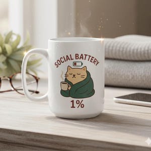 May include: White ceramic mug with the text "SOCIAL BATTERY" and a cartoon cat wrapped in a green blanket, holding a coffee cup. The mug also features the text "1%" and a low battery icon.