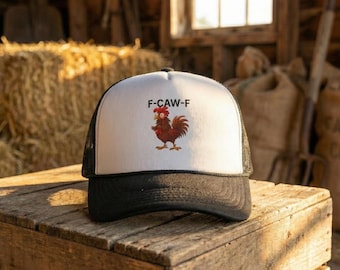 May include: A black and white trucker hat with a white front panel featuring a cartoon rooster and the text "F-CAW-F". The hat has a curved brim and a mesh back, sitting on a wooden surface.