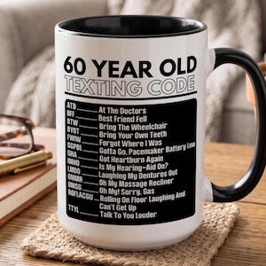 May include: A white ceramic mug with a black interior and handle, featuring the black text 60 YEAR OLD TEXTING CODE followed by a list of humorous acronyms and their meanings, such as ATD for At The Doctor's and TTYL for Talk To You Louder.