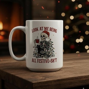 May include: White ceramic mug with a humorous Christmas design. The mug features a skeleton wearing a Santa hat, holding a cup, and wrapped in Christmas lights, with the text "LOOK AT ME BEING ALL FESTIVE & SH*T".