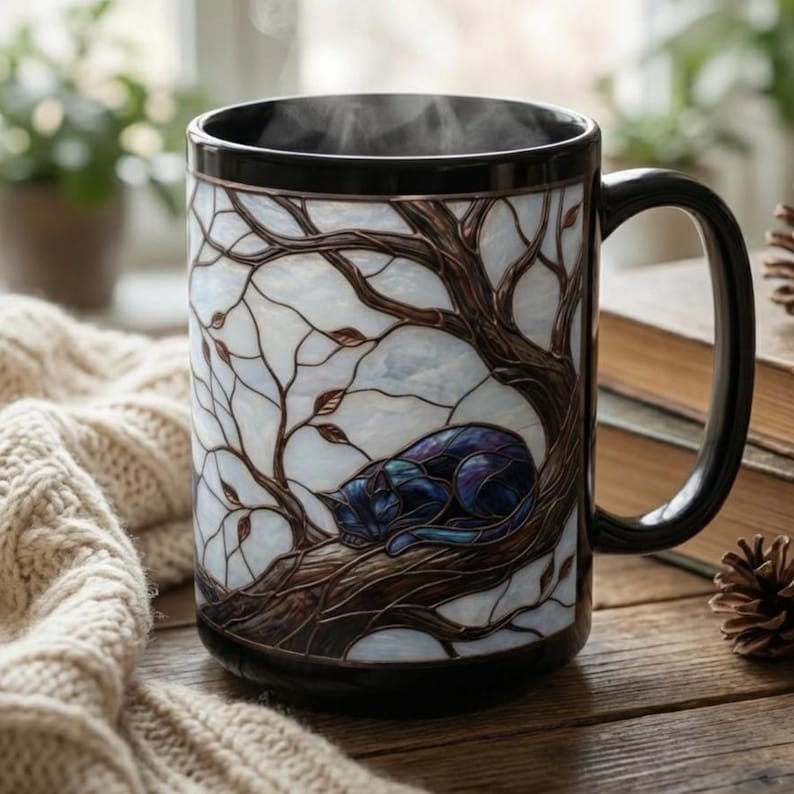 May include: A black ceramic mug with a stained-glass design featuring a sleeping cat on a tree branch. The mug has a black handle and rim, with steam rising from the contents. The background includes a cozy sweater and books.