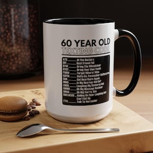 May include: White ceramic mug with a black interior and handle. The mug features the text "60 YEAR OLD TEXTING CODE" and a list of texting abbreviations. A spoon and a pastry are on a wooden surface.