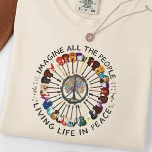 Op de afbeelding: Crèmekleurige sweatshirt met een cirkelvormige afbeelding met gitaren, muzieknoten en de tekst "IMAGINE ALL THE PEOPLE LIVING LIFE IN PEACE". Een zilveren ketting met een hanger is ook zichtbaar.
