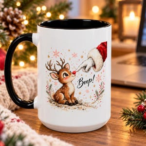 May include: A white ceramic mug with a black handle and interior rim. The mug features a graphic of a reindeer with a red nose being poked by a gloved finger. The text Boop! is printed in black script next to the reindeer, surrounded by stars.