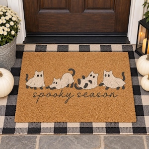Peut inclure: Un paillasson rectangulaire en fibre de coco beige avec quatre chats noirs portant des costumes de fantôme blancs. Le texte "spooky season" est imprimé en noir dans une police cursive sous les chats. Le tapis a une texture fibreuse.