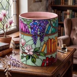 May include: A cylindrical ceramic vessel with a light pink interior rim and a stained glass-style exterior pattern featuring colorful books on shelves, purple wisteria blossoms, and light pink magnolia flowers, with visible steam rising from the top.