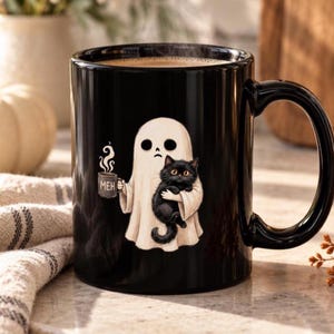 May include: A black ceramic mug featuring a graphic of a white ghost holding a small mug labeled MEH and cradling a fluffy black cat. The ghost has a simple face with two dark eyes and a straight mouth, while the cat has large orange eyes.