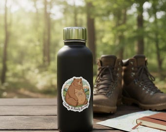 May include: A black insulated water bottle with a silver lid and a sticker featuring a cartoon bear. The bear is brown and sitting down with the text "Chunky & Ready for Naps". Hiking boots and a map are in the background.