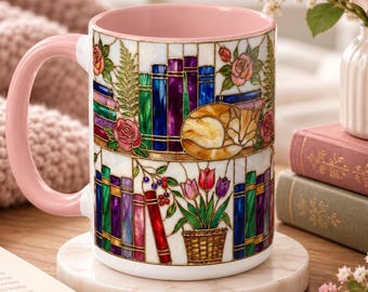 Op de afbeelding: Een keramische mok met een lichtroze handvat en binnenkant, voorzien van een rondlopende print in glas-in-loodstijl met boekenplanken, een slapende oranje kat, roze rozen en een mand met kleurrijke tulpen, inclusief blauwe, paarse en groene boeken.