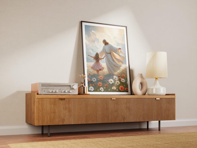 May include: A framed painting depicting a man in a white robe holding a young girl's hand as they walk through a field of flowers. The painting is displayed on a wooden cabinet with a stereo system and a lamp.