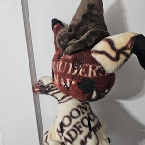 May include: A brown and white plush toy with a brown pointed hat. The toy has a black nose and eyes and is wearing a white robe with brown text.