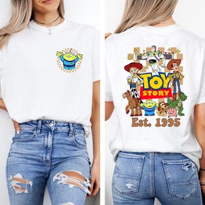 May include: White t-shirt featuring a cartoon alien on the front and a Toy Story graphic on the back. The back graphic includes characters like Woody, Buzz Lightyear, and Jessie, with the text "TOY STORY" and "Est. 1995".