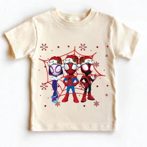 May include: Cream-colored t-shirt featuring three cartoon Spider-Man characters wearing Santa hats, set against a red spiderweb design with snowflakes. The characters are in their signature costumes. A festive holiday-themed graphic tee.